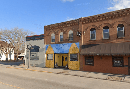 A colorful mural of mountains and corn adorns a brick building on a street with trees and shops in the background.