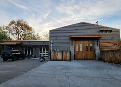 A rustic building with wooden doors and a gray exterior, featuring a parking area and outdoor seating.