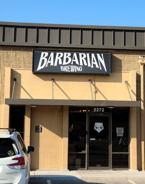 Sign for Barbarian Brewing on a building exterior, with a white vehicle parked in front. Clear blue sky above.