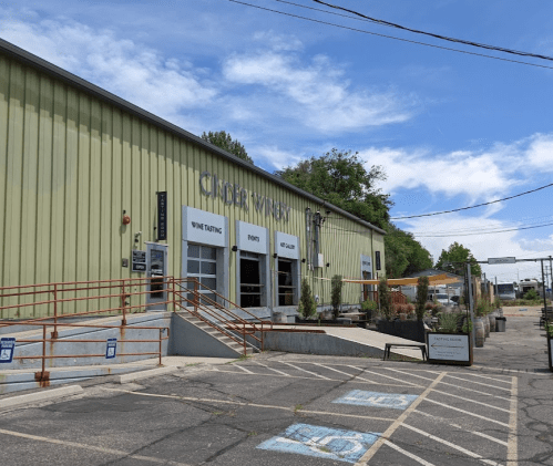 Exterior of a green winery building with a sign for wine tasting, featuring a patio and clear blue sky.