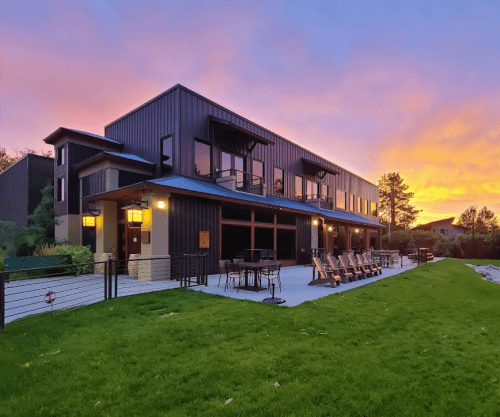 Modern building with large windows and outdoor seating, set against a vibrant sunset sky. Lush green lawn in foreground.