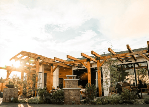 A modern restaurant with wooden beams, outdoor seating, and sunlight shining through, surrounded by greenery.