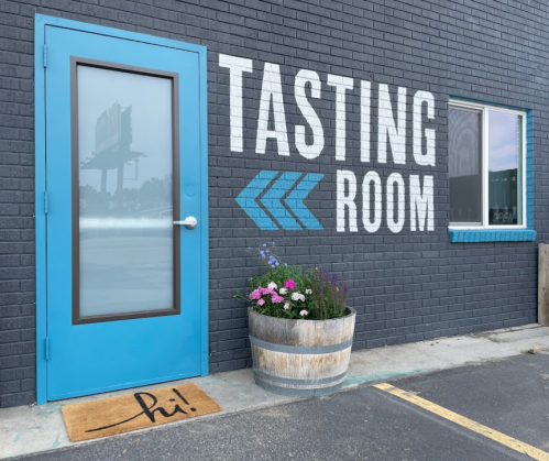 A blue door labeled "Tasting Room" next to a window, with a flower pot and a welcome mat that says "hi!" in front.