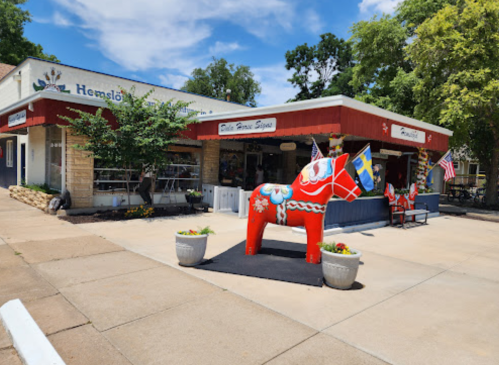 A colorful Dala horse statue stands outside a shop with flags and greenery in a sunny setting.