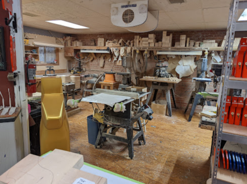 A woodworking shop filled with tools, wood pieces, and equipment, featuring a cluttered workbench and shelves.