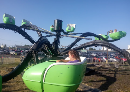 A girl sits in a green ride resembling an octopus, with other riders in the background and cars parked nearby.