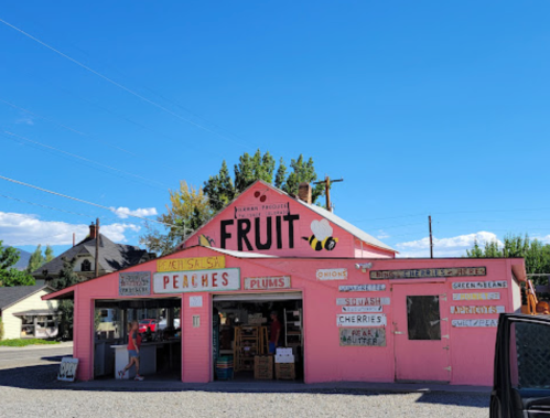 A pink fruit stand with signs for peaches, plums, and other produce under a clear blue sky.