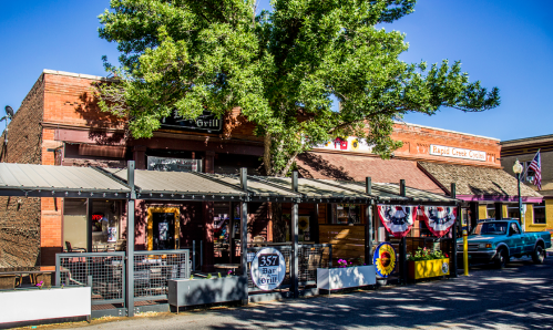A rustic bar and grill with outdoor seating, surrounded by trees and colorful banners in a small town setting.
