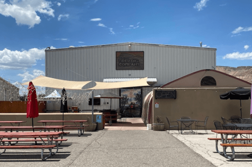 Exterior of a brewing company with outdoor seating, a large building, and a clear blue sky.