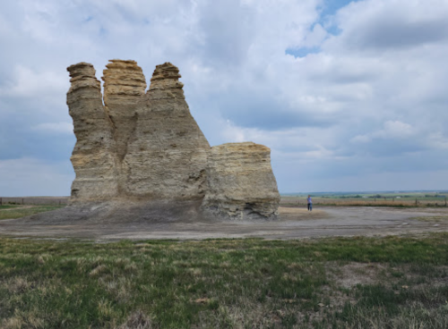 A person stands next to a large rock formation with three distinct peaks under a cloudy sky.