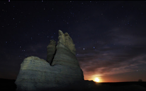 A rocky formation silhouetted against a starry night sky with a glowing horizon at sunset.
