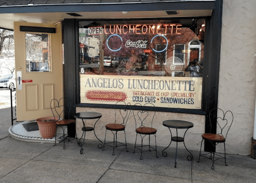 Exterior of Angelo's Luncheonette with a sign, neon lights, and empty chairs outside.