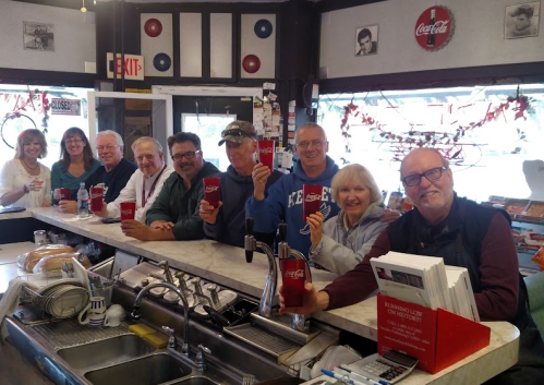 A group of nine people smiling and holding Coca-Cola cups at a diner counter, with a retro decor in the background.