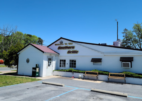 A white building with a maroon roof, sign reading "Welcome To The Southern Grille," and benches outside. Clear blue sky.