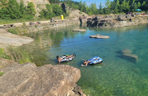 Two people float on inner tubes in a clear blue water area surrounded by rocks and greenery.