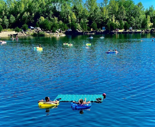Two people relax on colorful floats in a serene lake, surrounded by others enjoying the water on a sunny day.