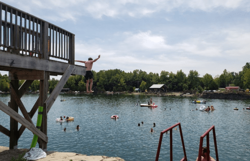 A person jumps off a diving board into a lake, surrounded by swimmers and inflatable floats on a sunny day.