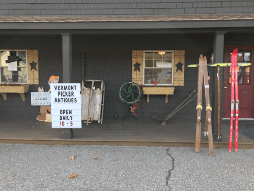 A porch displaying a sign for Vermont Picker Antiques, vintage skis, and various antique items. Open daily 10-5.