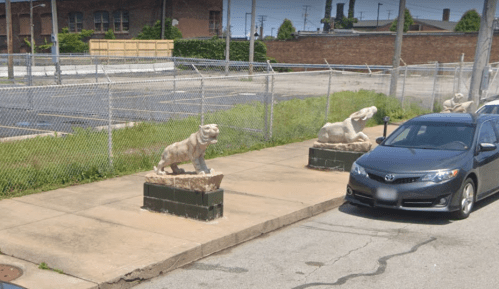 Two stone animal sculptures on a sidewalk beside a parking lot, with a car parked nearby and a chain-link fence in the background.