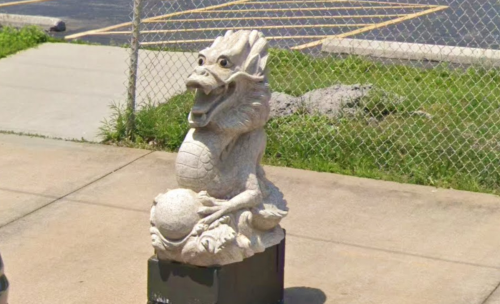 A stone dragon statue sitting on a pedestal, holding a sphere, with a grassy area and chain-link fence in the background.