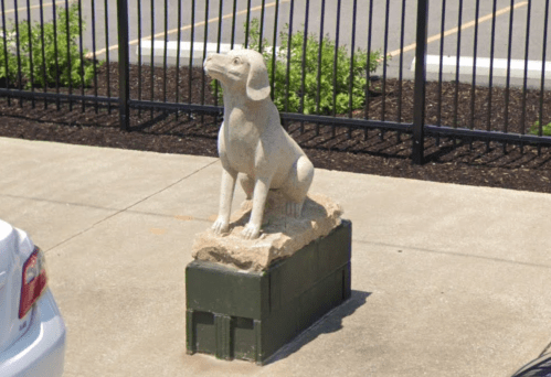 A stone statue of a sitting dog on a rock pedestal, placed on a concrete surface near a parking area.