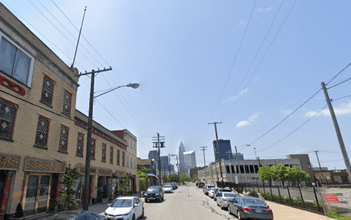 A city street lined with parked cars, featuring buildings and a skyline in the background under a clear blue sky.