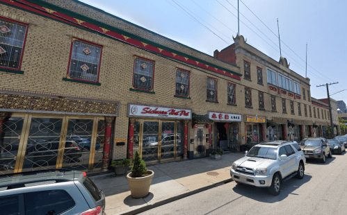 A street view of a brick building with decorative windows, featuring shops and parked cars along the sidewalk.
