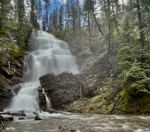 A serene waterfall cascades over rocky terrain, surrounded by lush green trees and a clear blue sky.