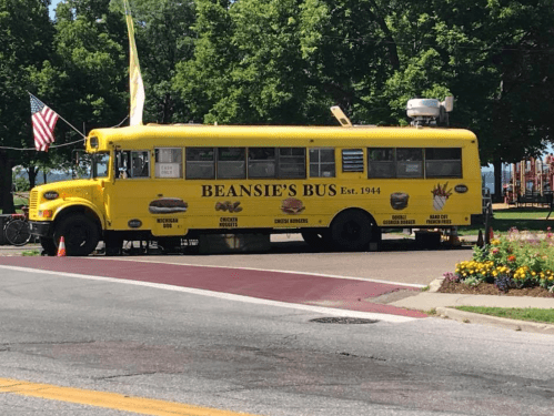 A bright yellow food bus named "Beansie's Bus" parked near a park, featuring various menu items and an American flag.