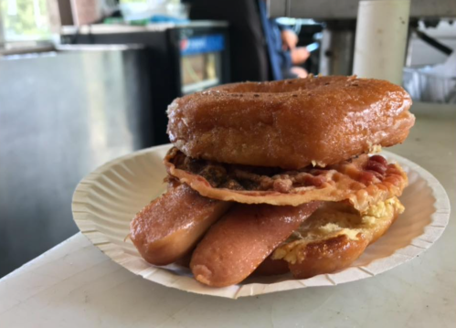 A stacked sandwich featuring a glazed donut, bacon, sausage, and a fried egg on a paper plate.