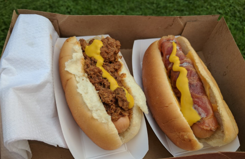 Two hot dogs in a cardboard box: one topped with chili and mustard, the other with bacon and mustard.