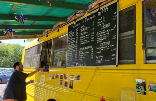 A man orders food at a yellow food truck with a large menu board displaying various items and prices.