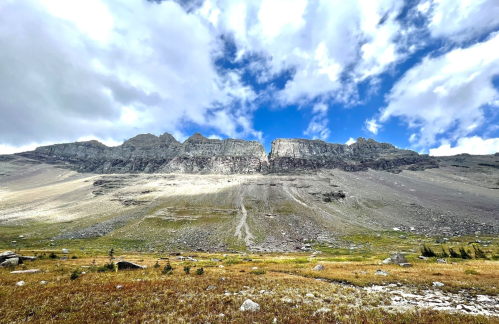 A dramatic mountain range under a blue sky with scattered clouds, featuring rocky cliffs and a grassy foreground.
