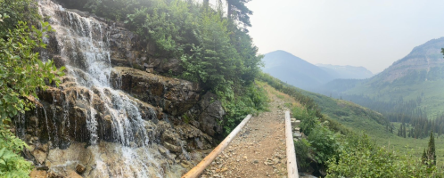 A scenic view of a waterfall beside a dirt path, surrounded by lush greenery and misty mountains in the background.