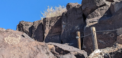 Rocky landscape with petroglyphs on a cliff, wooden posts, and sparse vegetation against a clear blue sky.