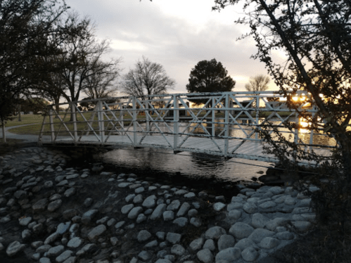 A white bridge spans a calm waterway, surrounded by trees and rocks, with a sunset in the background.