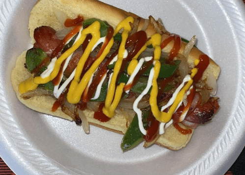A hot dog in a bun topped with grilled onions, green peppers, ketchup, mustard, and mayonnaise on a paper plate.