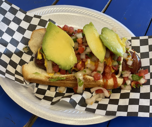 A hot dog in a bun topped with avocado, diced tomatoes, onions, mustard, and ketchup, served on a checkered paper.