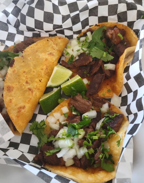 Three tacos filled with beef, topped with onions and cilantro, served with lime wedges in a checkered paper basket.