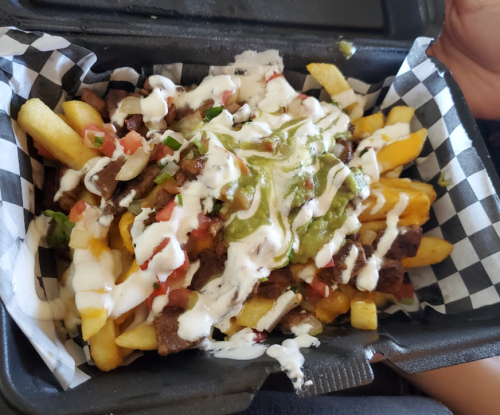 A takeout container filled with loaded fries topped with beef, guacamole, diced tomatoes, and drizzled with sauce.