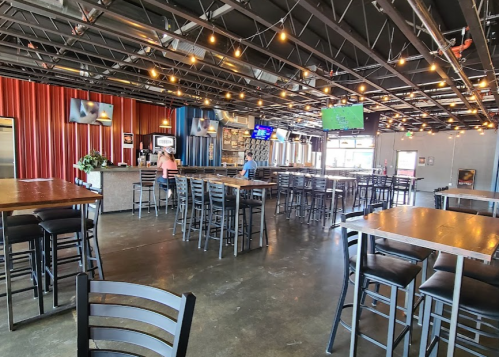 A spacious restaurant interior with high tables, bar stools, and string lights, featuring a bar and TVs on the walls.