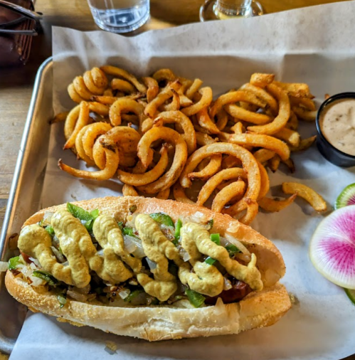 A hot dog topped with mustard and onions, served with a side of curly fries and a small cup of dipping sauce.