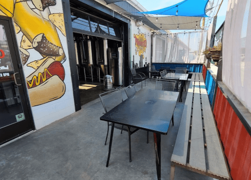 Outdoor seating area with metal tables and benches, colorful walls, and a brewery visible in the background.
