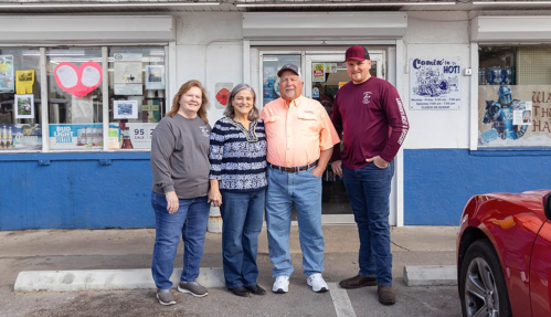 Four people stand outside a store, smiling, with a blue and white building and windows displaying various signs behind them.