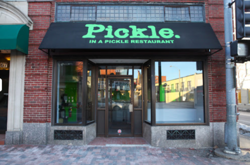 Exterior of "Pickle. in a Pickle" restaurant, featuring a black awning with green text and large glass windows.