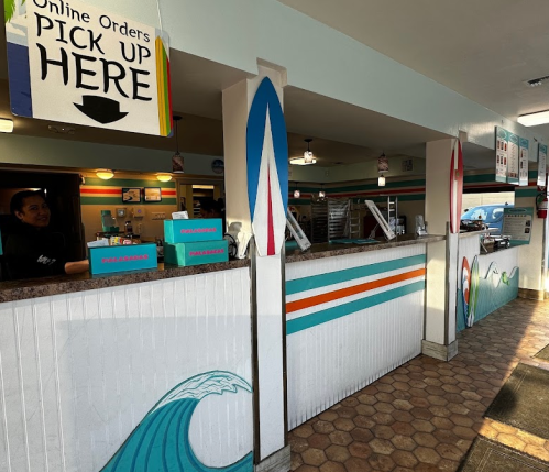 A colorful counter with surfboard decor, featuring a sign for online order pickup and a friendly staff member inside.