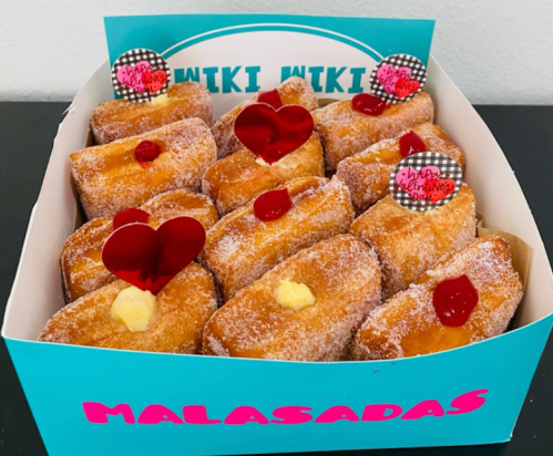 A box of malasadas, filled with cream, topped with red hearts and festive decorations, on a colorful background.
