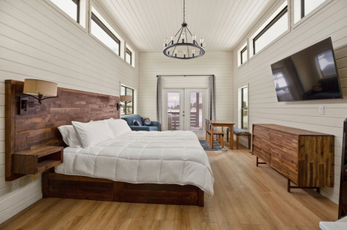 Cozy bedroom with a wooden bed, large windows, a TV, and a seating area, featuring modern decor and warm tones.