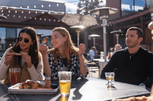 Three people enjoying food and drinks at an outdoor table, with warm lighting and a casual atmosphere.