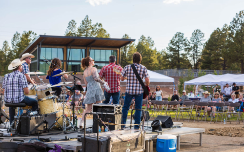 A band performs on stage outdoors, with an audience seated in the background and trees and a modern building behind them.